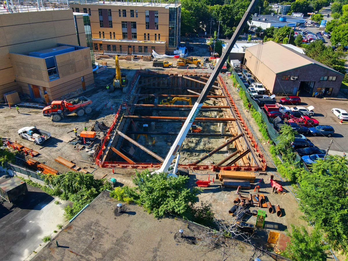 Aerial view of a deep urban construction excavation site with steel cross-bracing, yellow excavators, and a crane, adjacent to a multi-story brick building.